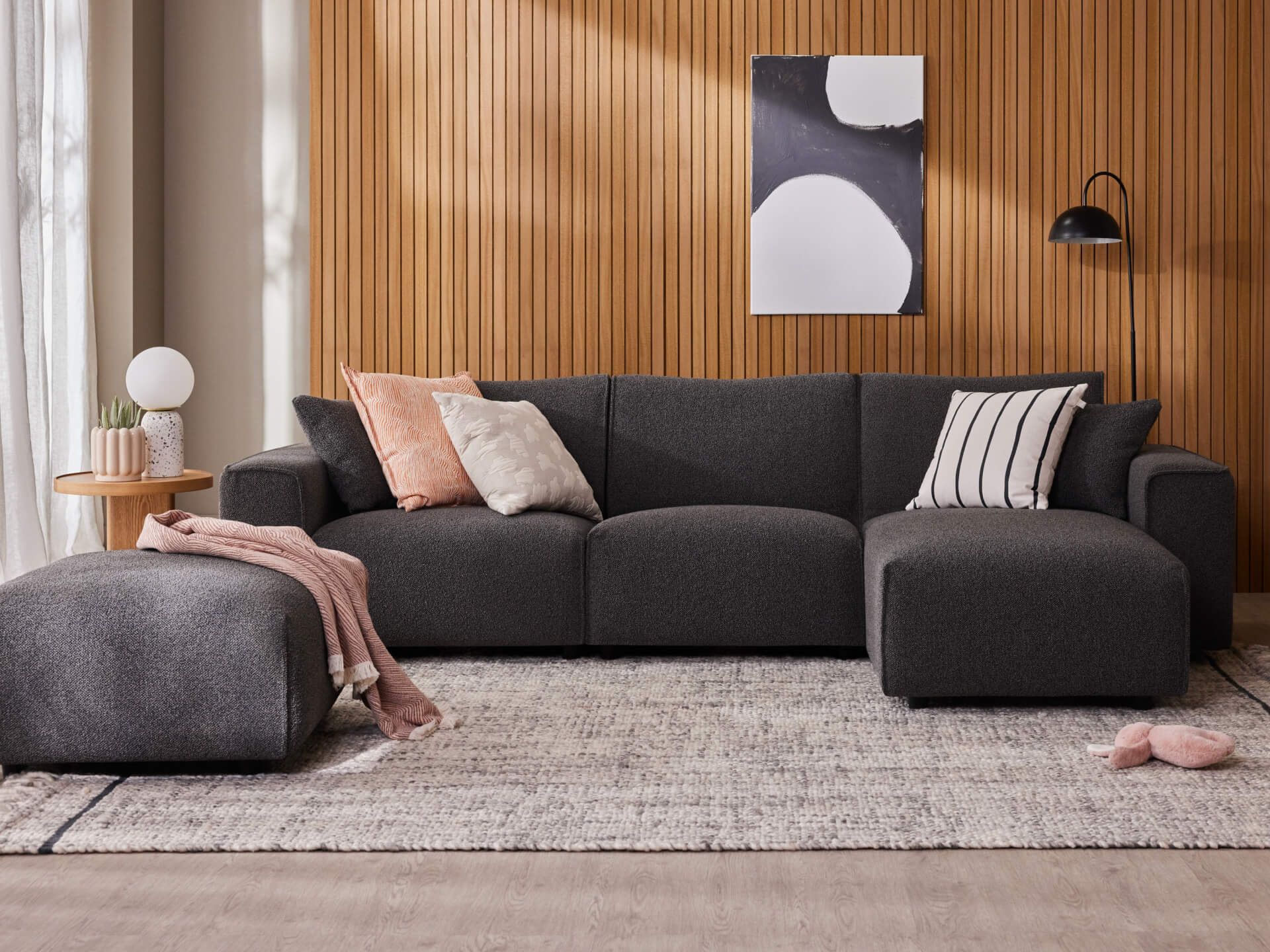Dark grey sectional sofa on a light rug, wooden panel wall, abstract art, cushions, side table with lamp and plant, pouffe and throw blanket.
