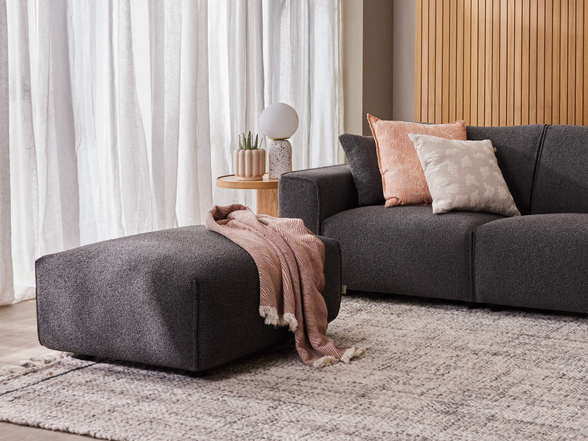 Modern living room with a dark grey sofa and ottoman, pink throw, and decorative cushions. Neutral rug and wooden accents complete the cosy setting.