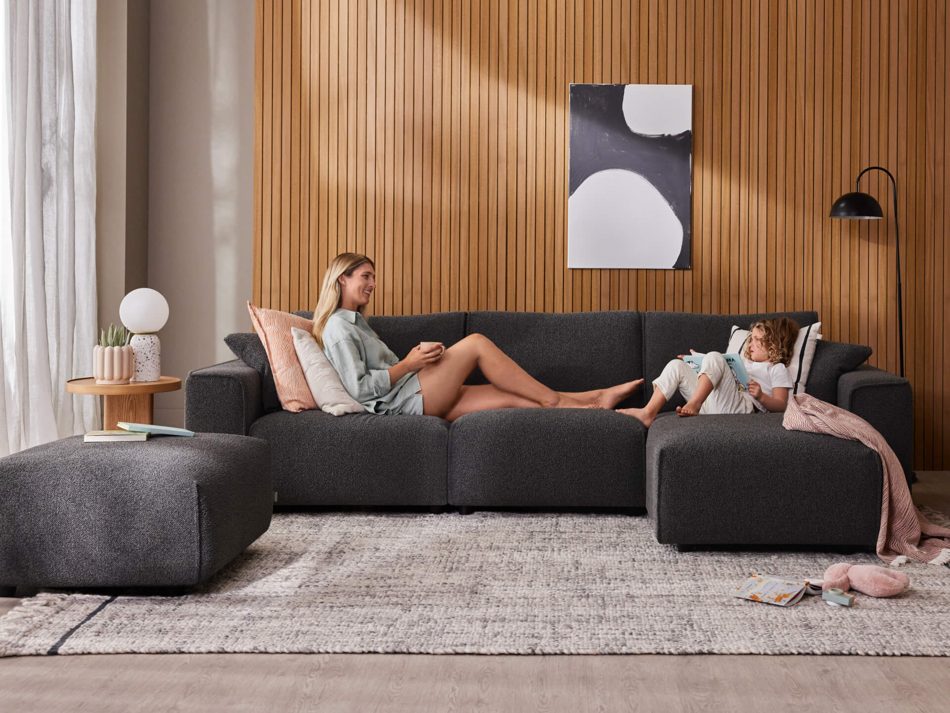 Modern lounge room with dark grey sofa, light rug, round side table, and ottoman. Wood panel wall, abstract art, and floor lamp add style.