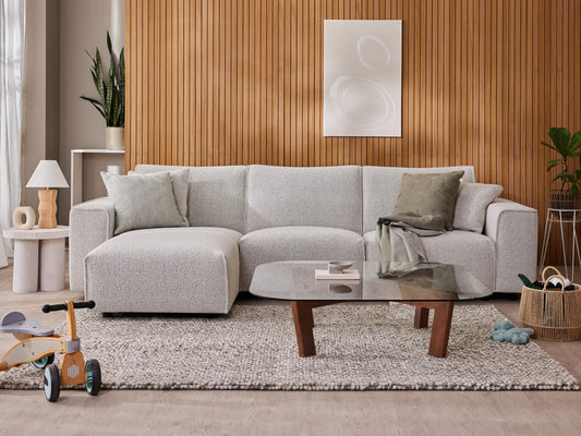 Light grey L-shaped sofa with two green cushions, wooden panelled wall, abstract art, glass coffee table, grey rug, and decor accents.