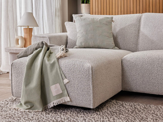 Light grey textured corner sofa with a soft green throw and a grey cushion. Nearby is a round side table with a white lamp. Neutral decor setting.