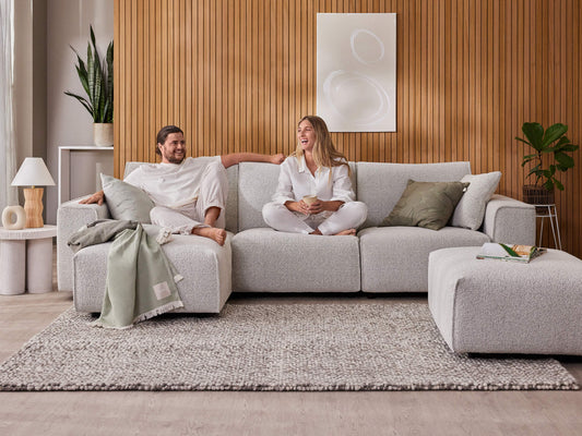 Light grey sofa with plush cushions on a textured rug. Wooden panel wall, decorative plants, and minimalist art complete the cosy living room setting.