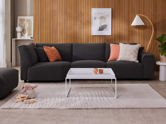 Modern living room with dark grey sofa, orange and beige cushions, white square coffee table, wooden panelled wall, and light grey rug.
