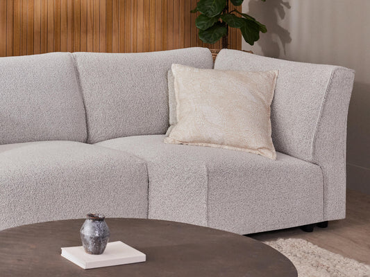 Curved light grey sofa with beige cushion, wooden panel wall, round brown table with a small vase, and a potted plant in the background.