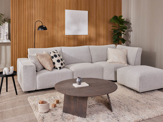 Modern living room with a light grey sectional sofa, round dark wood coffee table, and a textured rug. Neutral decor and wooden panel wall.