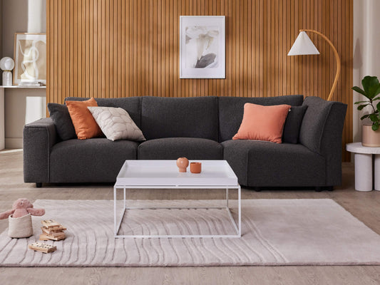Dark grey sectional sofa with orange and beige cushions on a light rug. White coffee table, wood panel wall, indoor plant, floor lamp, and abstract art.