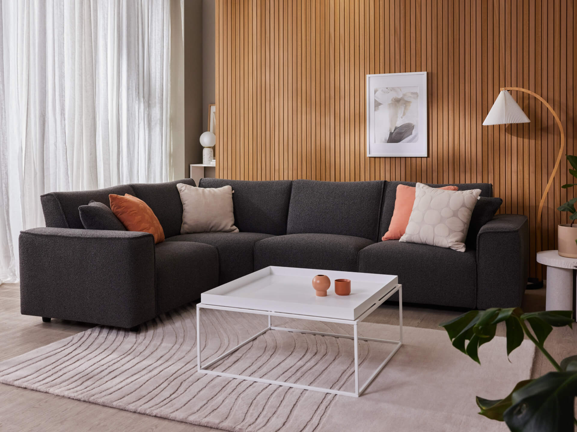 Dark grey sectional sofa with mixed cushions in a modern living room. White square coffee table, wood-panelled wall, and framed art above.