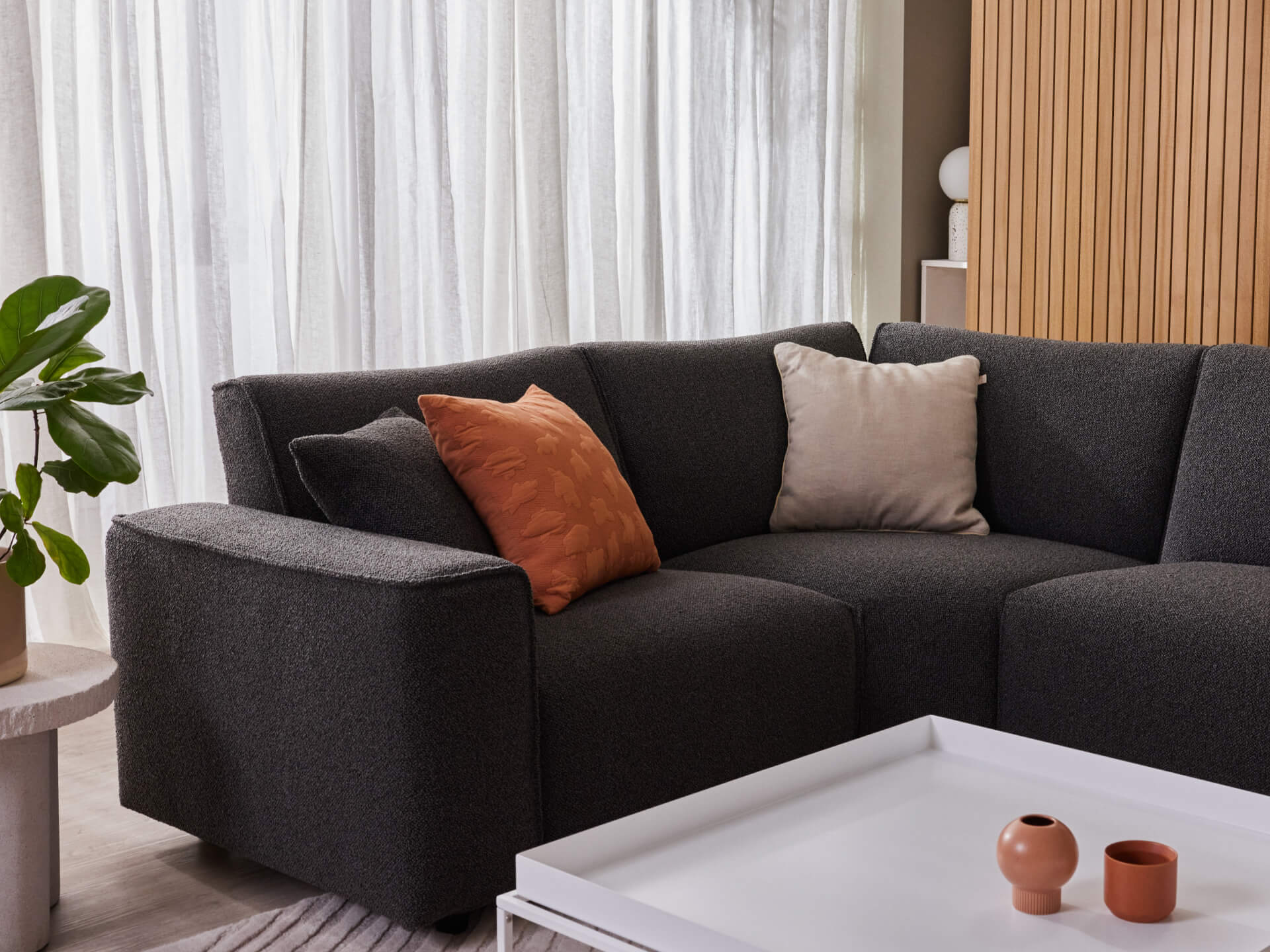 Dark grey sectional sofa with orange and cream cushions, set in a modern room with white curtains, wooden panel wall, and white coffee table.