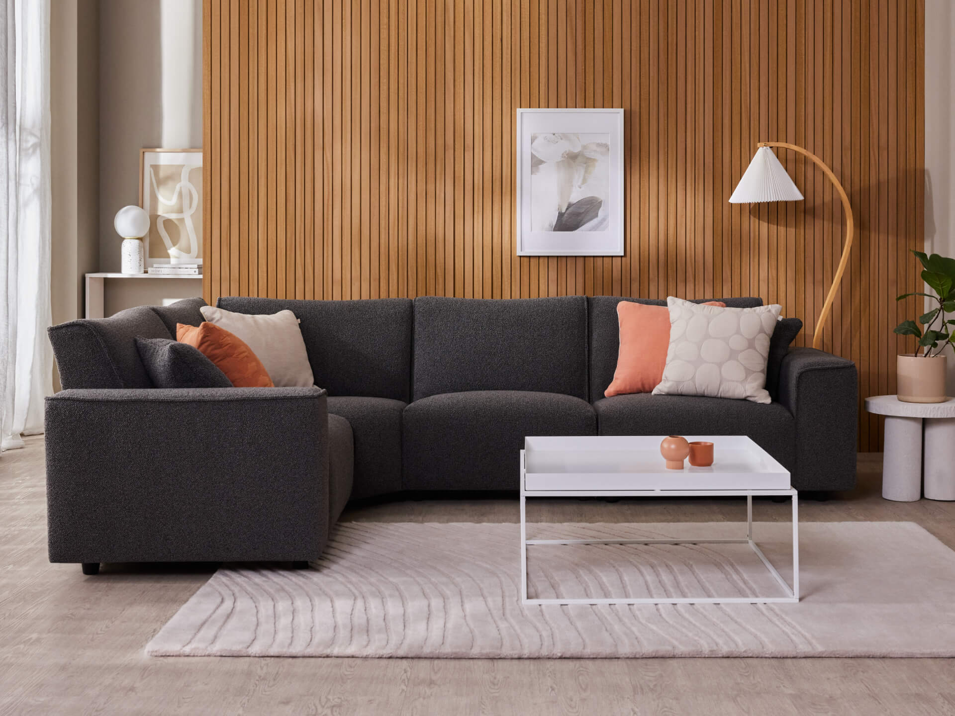 Dark grey modular sofa with assorted cushions against a wooden panelled wall. White coffee table on a light grey rug; modern lamp and decor.