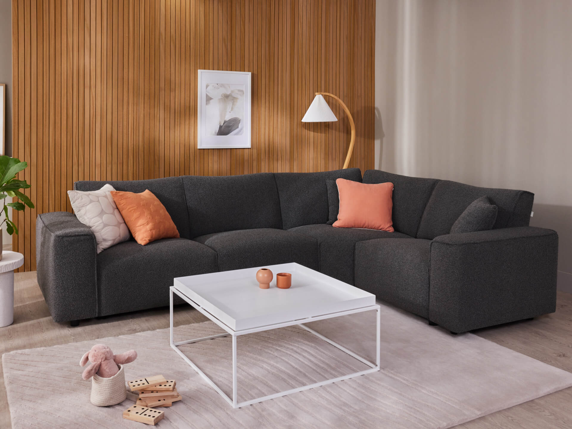 Dark grey sectional sofa with peach cushions, white square coffee table, on beige rug. Wooden panel wall, floor lamp, and plant in background.