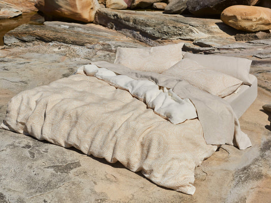 Bedding set with beige and cream patterns on a bed, placed outdoors on a rocky surface. Light-coloured pillows and sheets.