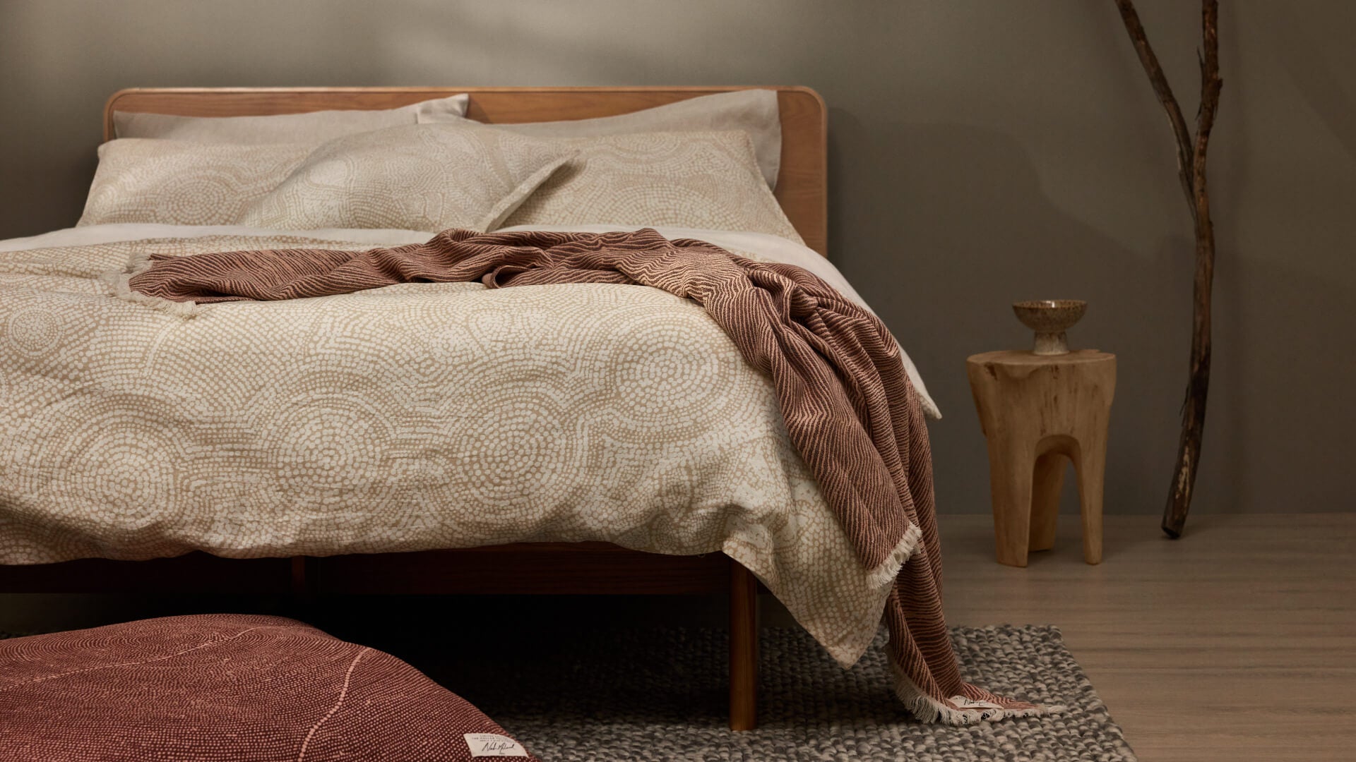 Minimalist bedroom with a wooden bed, light brown patterned bedding, and a brown blanket. A small wooden side table and grey rug complete the look.