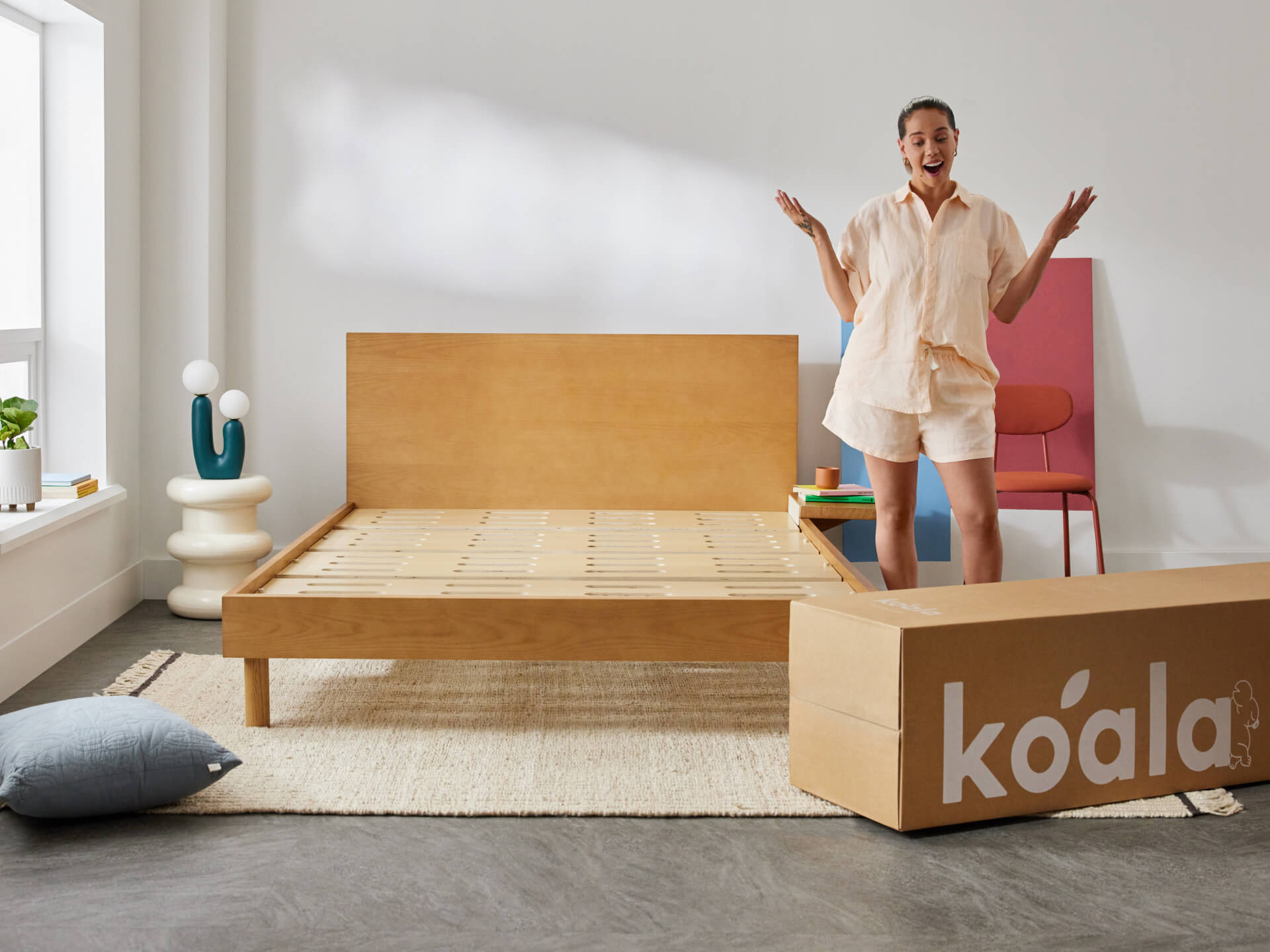Woman excitedly stands next to an assembled wooden bed frame. A large cardboard box labeled "koala" is on the floor. Minimalist room decor.