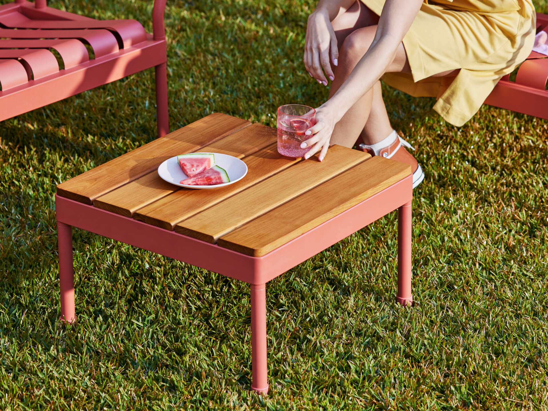 Outdoor wooden coffee table with a pink metal frame on grass. A woman in a yellow dress holds a drink. Plate with watermelon slices on table.