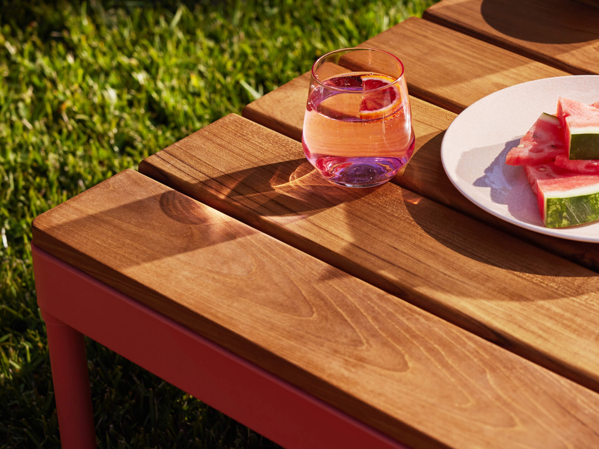 Wooden outdoor table with a red frame, featuring a glass of pink drink and a plate of watermelon slices, set on green grass.