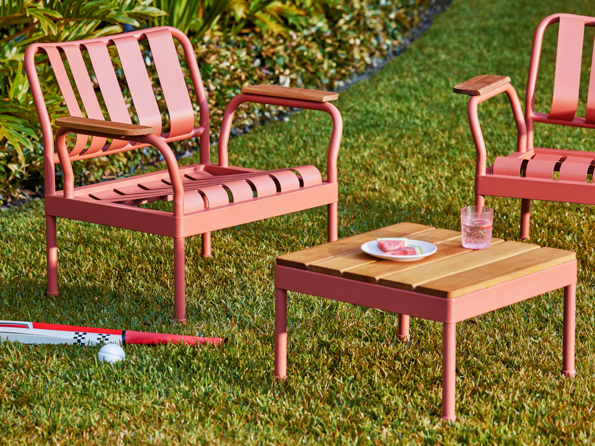 Outdoor setting with two coral pink metal chairs and a matching wooden table on grass. A cricket bat and ball lie nearby. A drink and plate on the table.