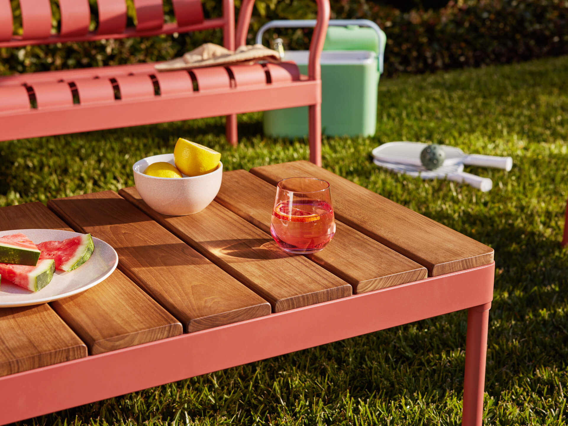 Outdoor setting with a pink metal and wood table, holding a drink and lemons. Background includes a pink chair, cooler, and tennis racquets on grass.