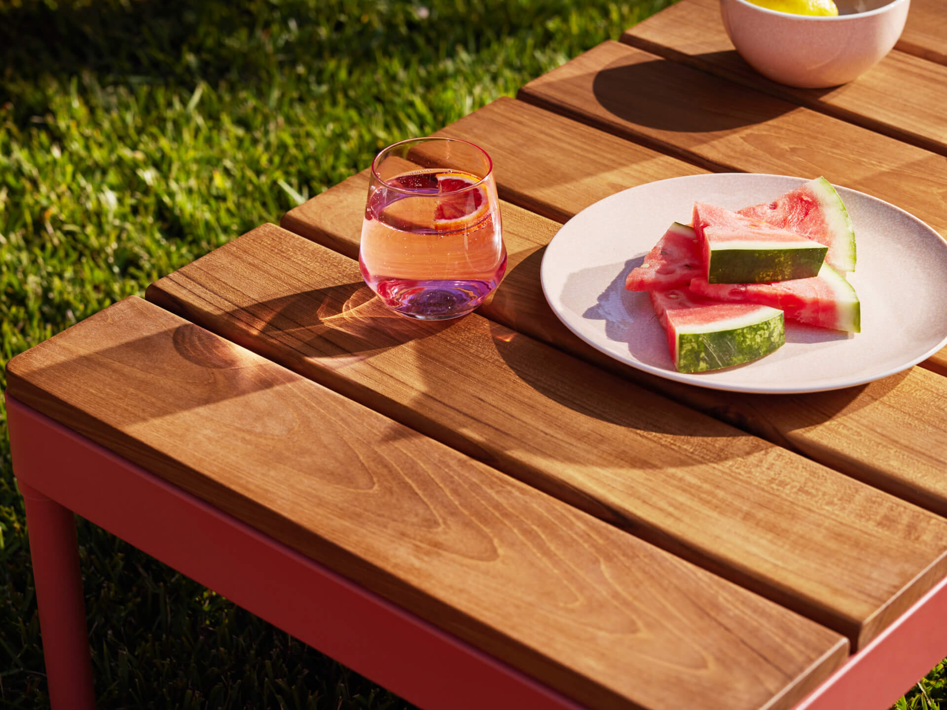 Wooden outdoor table with red frame, holding a glass of pink drink and a plate with watermelon slices. Green grass in the background.