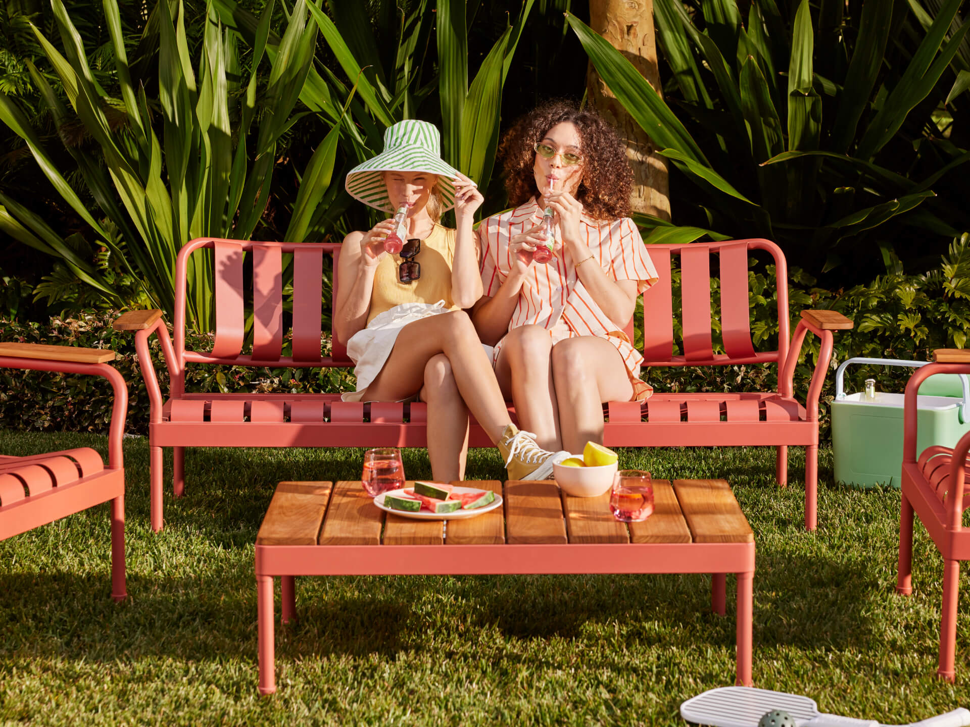 Two people sit on a coral outdoor bench with drinks. A matching coffee table holds watermelon, bowl, and glasses. Lush greenery in the background.
