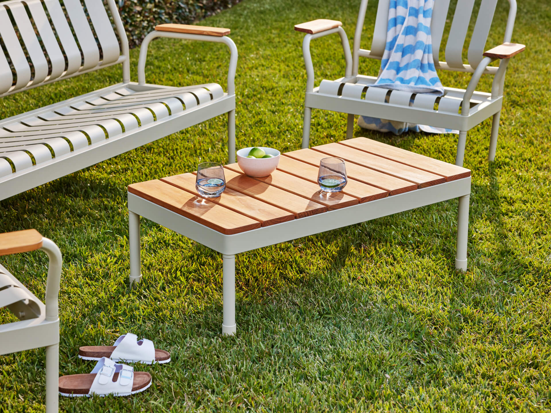 Outdoor setting with a beige metal and wood table, two glasses, a bowl, and surrounding chairs on green grass. White sandals nearby.
