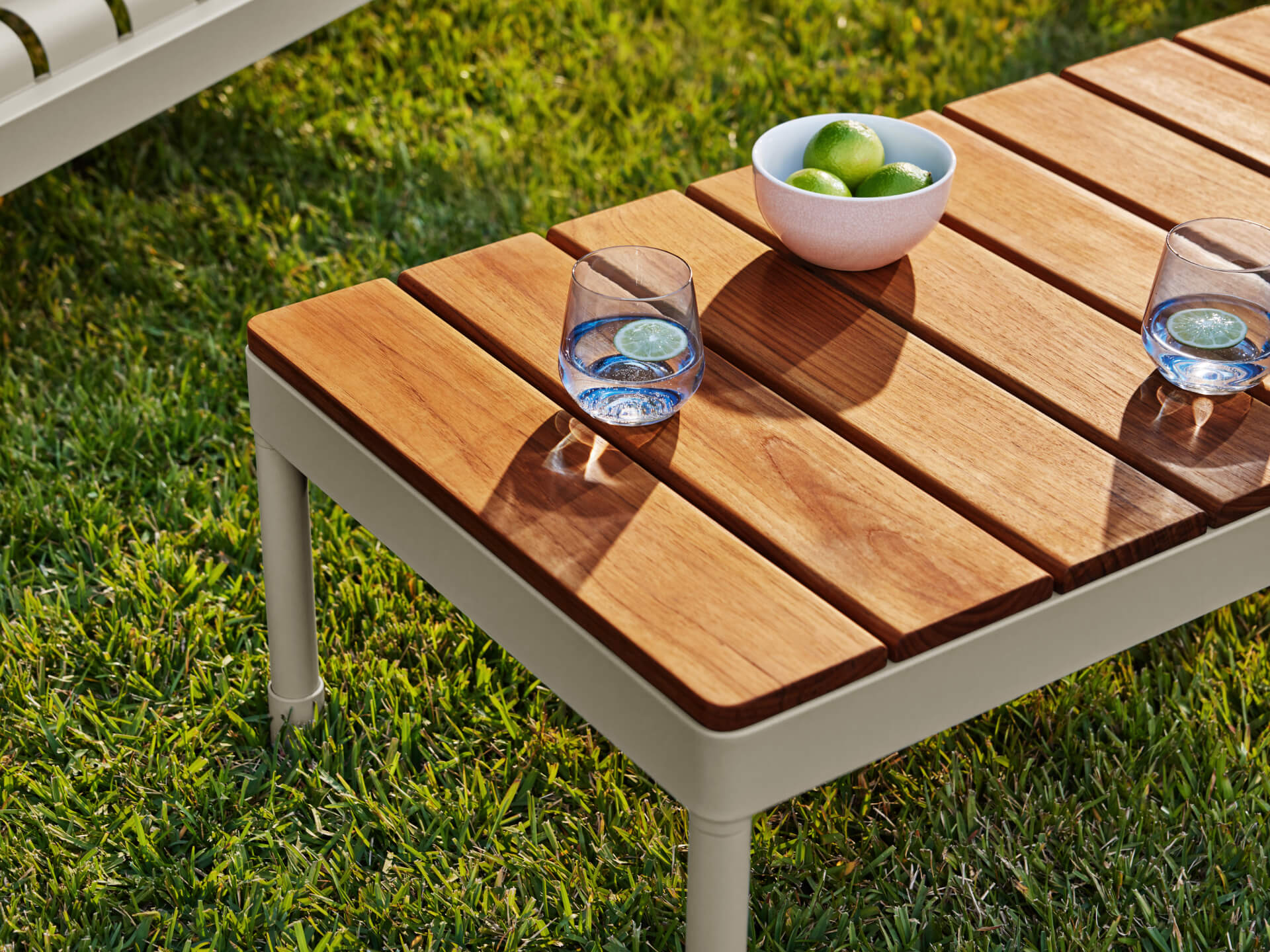 Wooden outdoor coffee table on grass with two glasses of water and a bowl of green fruit. Beige metal legs. Shadows cast on table surface.