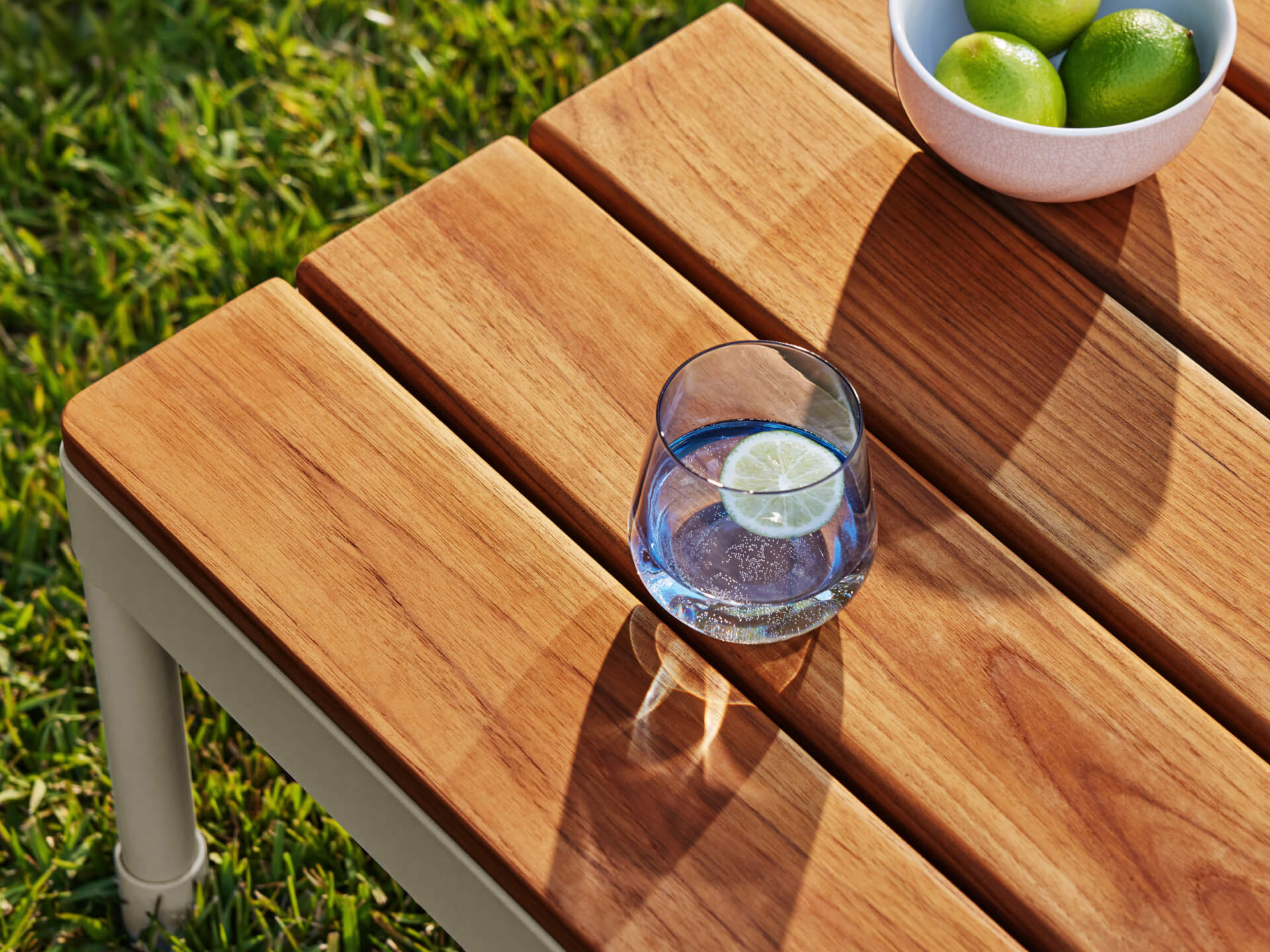 Wooden outdoor table with metal legs on grass, featuring a glass of water with a lime slice and a bowl of green lemons.