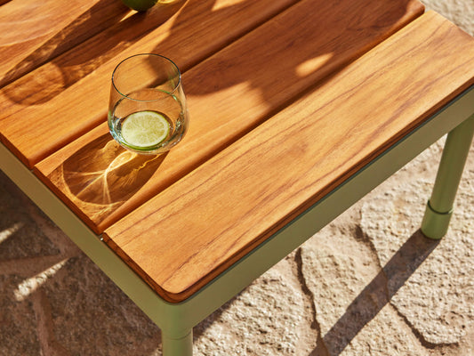 Wooden outdoor table with a light brown top on a light green frame. A glass of water with a lime slice sits on it, casting a shadow. Stone flooring.