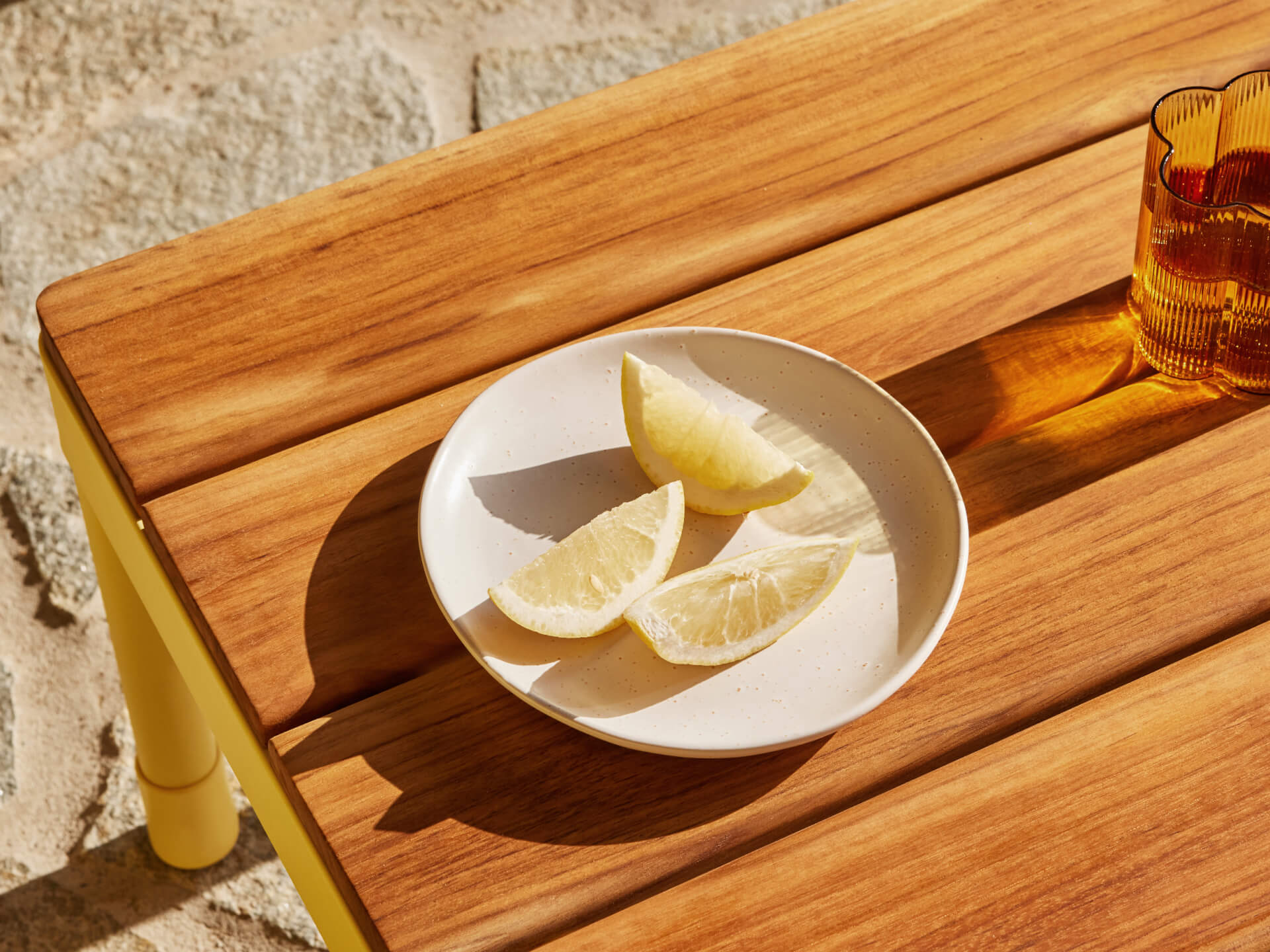 Wooden table with a small white plate of lemon wedges and a textured amber glass. Sunlit stone floor in the background.