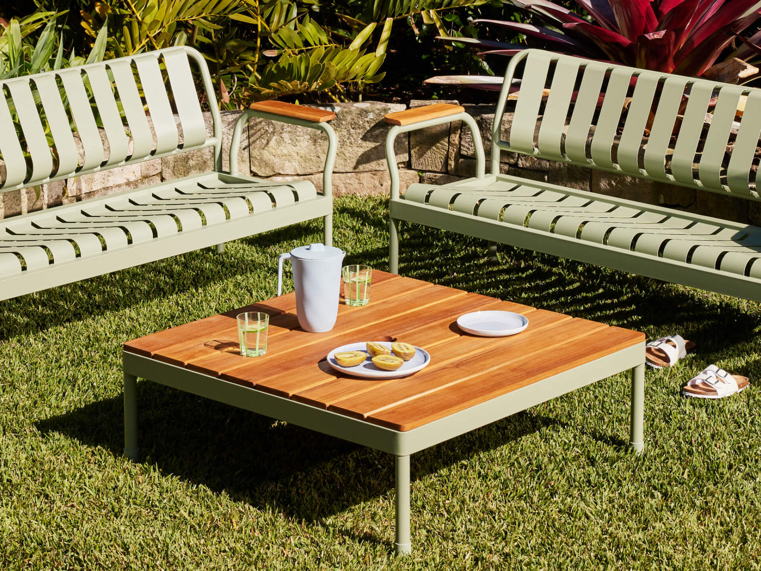 Outdoor setting with two light green benches and a wooden coffee table on grass. Table holds a jug, glasses, and plates. Lush garden backdrop.