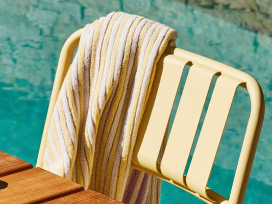 Yellow metal chair by a pool, draped with a striped towel. Wooden table edge in the foreground. Sunny day by the water.