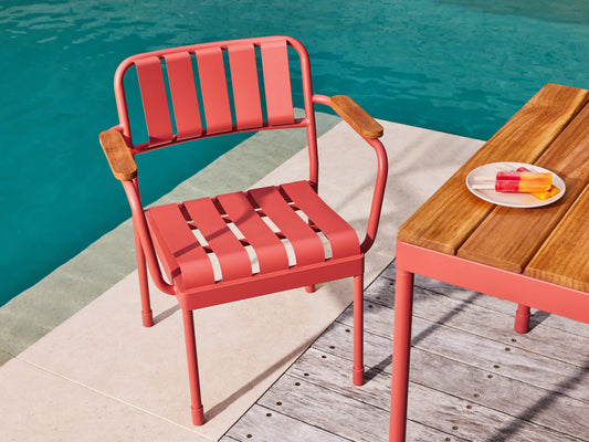 Coral chair with wooden armrests beside a wooden table. Plate of icy poles on table. Image is set by a poolside on a sunny day.