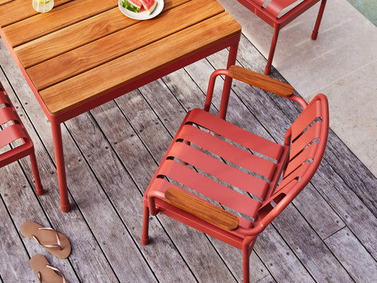 Red metal patio chair and table with wooden slats on a weathered deck. Casual sandals are placed nearby, and a plate of fruit is on the table.