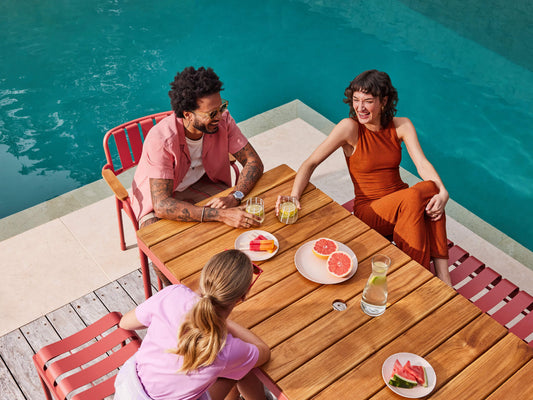 Three people sit at a wooden outdoor table with red chairs by a pool, enjoying drinks and fruit plates. The setting is vibrant and relaxing.