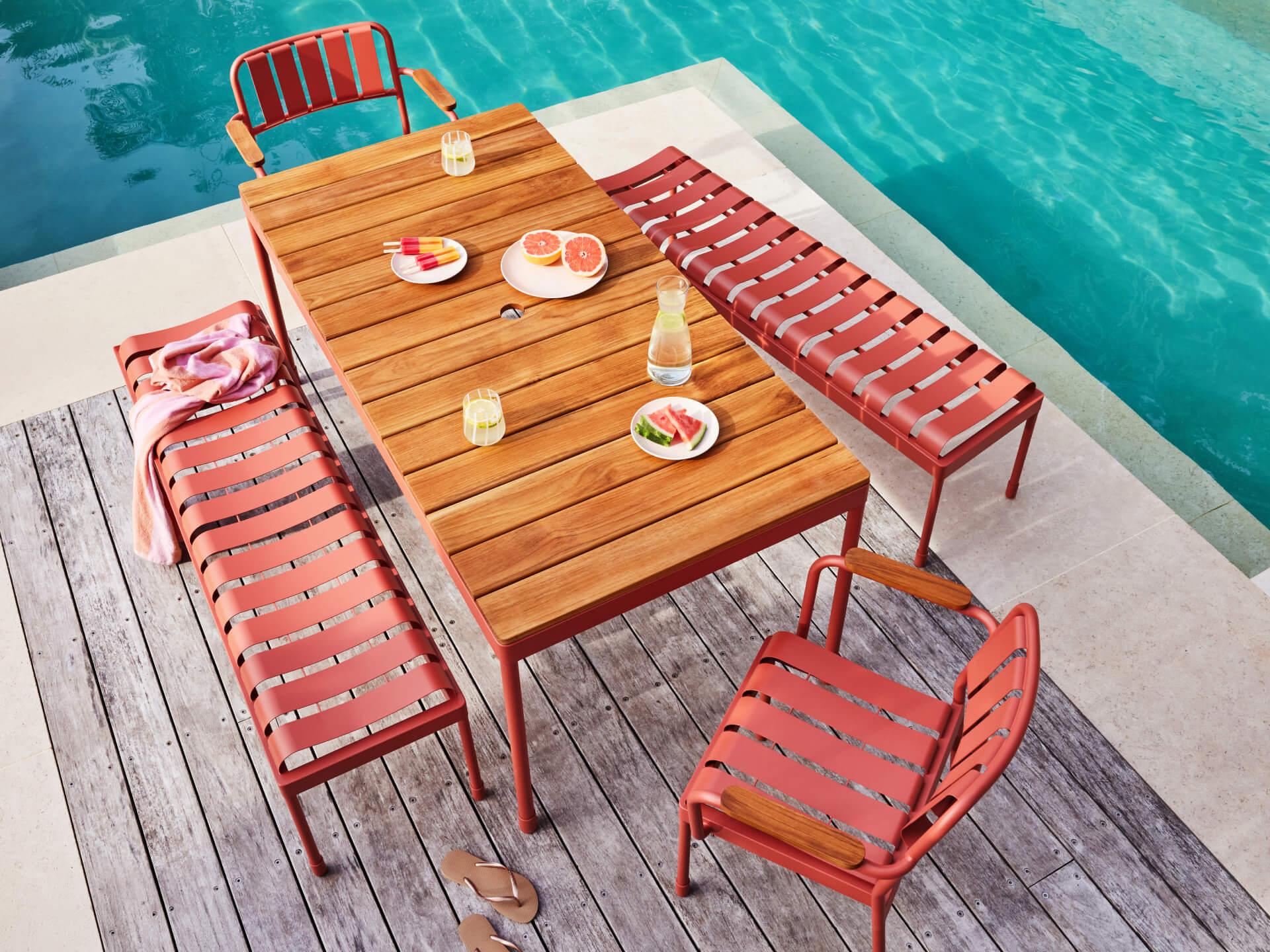 Outdoor setting with a wooden table, red metal chairs, and benches by a pool. Drinks and fruit are on the table, and a towel is draped over a bench.