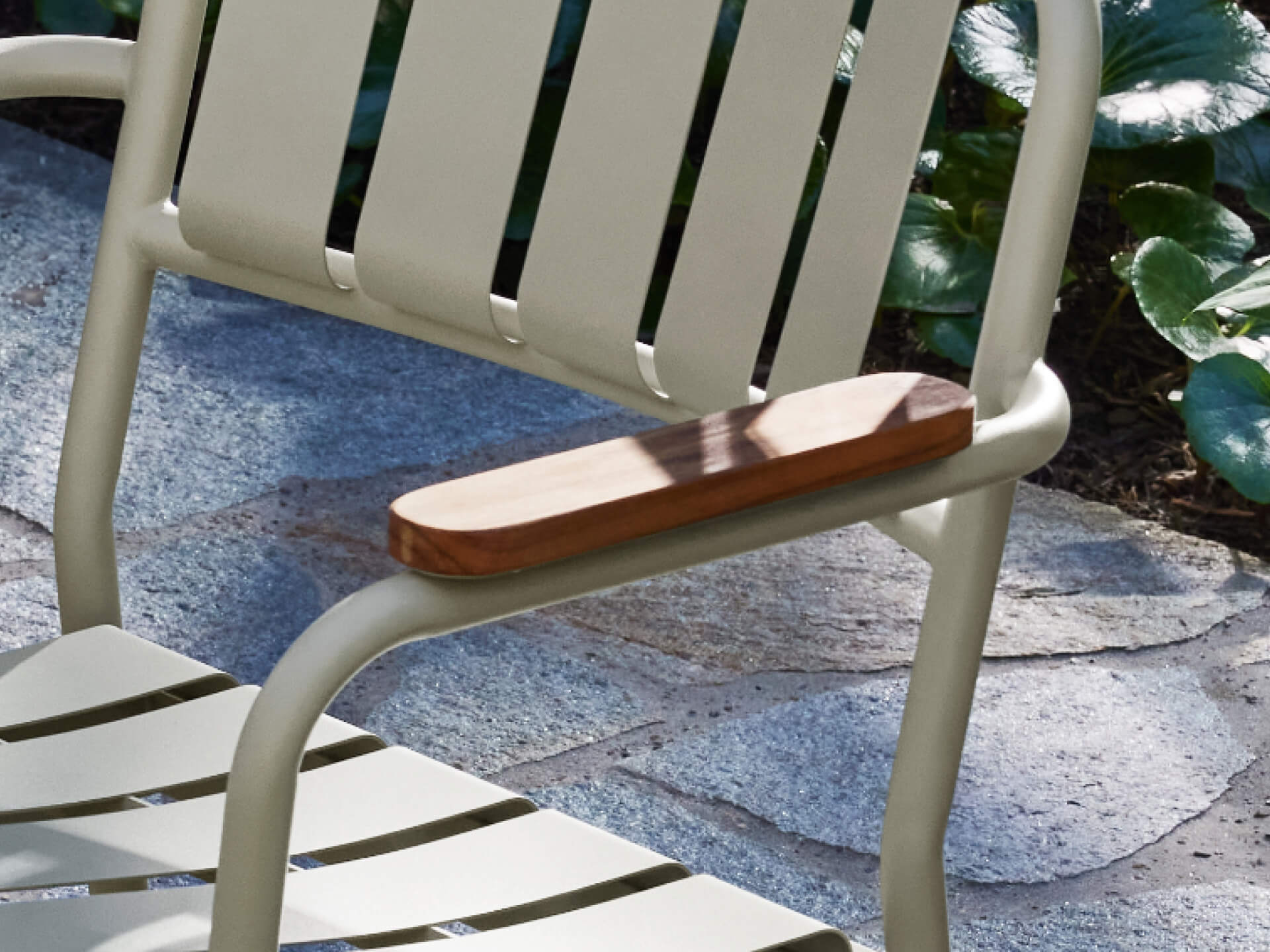 Outdoor metal chair with light green slats and a wooden armrest detail, set on a stone patio with green foliage in the background.