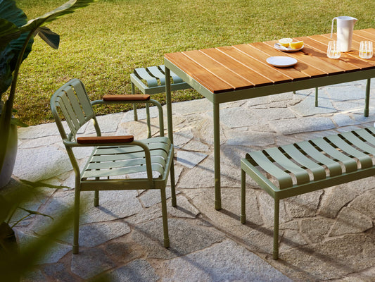 Garden setting with a wooden table and green metal chairs and bench on a stone patio, overlooking a grassy lawn. Glasses and plates on the table.