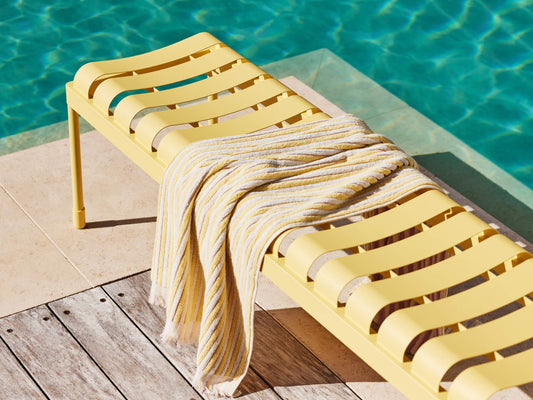 Yellow slatted outdoor bench beside a pool, with a striped yellow and white towel draped over it, set on a wooden deck.