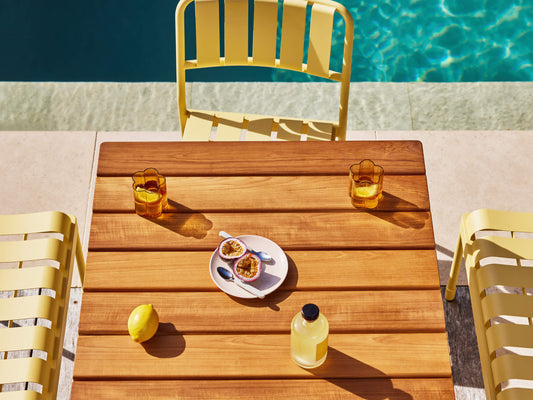 Wooden outdoor table with yellow chairs by a pool. On the table are two amber glasses, a lemon, a drink bottle, and a plate with passionfruit halves.