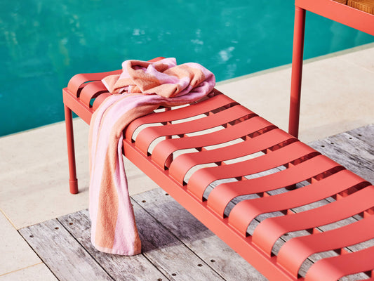Red outdoor bench with slatted design by a pool, draped with a pink towel. Set on light stone tiles with wood planks nearby.