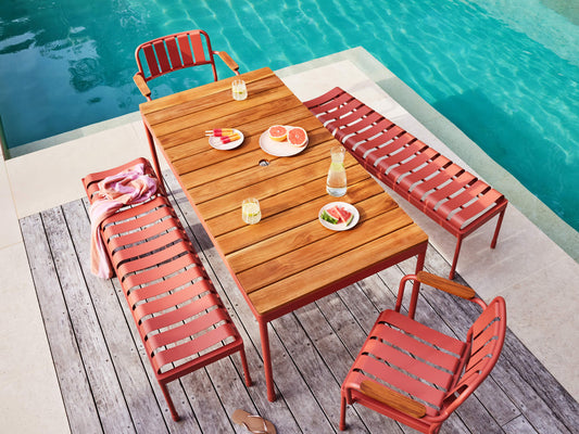 Outdoor setting with a wooden table and red metal chairs on a deck beside a pool. Table is set with juice, sliced fruit, and glasses.