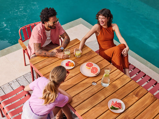 Three people relax around a wooden outdoor table with drinks and fruit plates, next to a pool. Red chairs add a pop of colour.