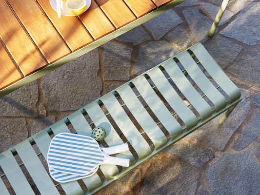 Outdoor setting with a wooden table and green metal bench on stone paving. Bench holds a striped paddle and ball.