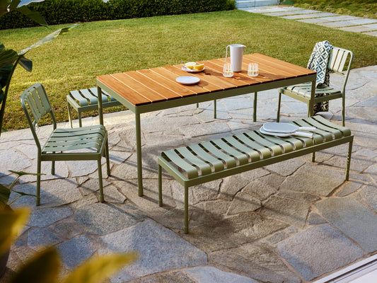 Outdoor setting with a wooden table, two green chairs, and a matching bench on a stone patio. Plates, glasses, and a jug adorn the table.