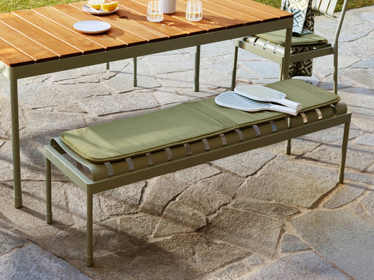 Outdoor setting with a green cushioned bench and wooden-top table on stone pavement. Nearby is a striped cushion.