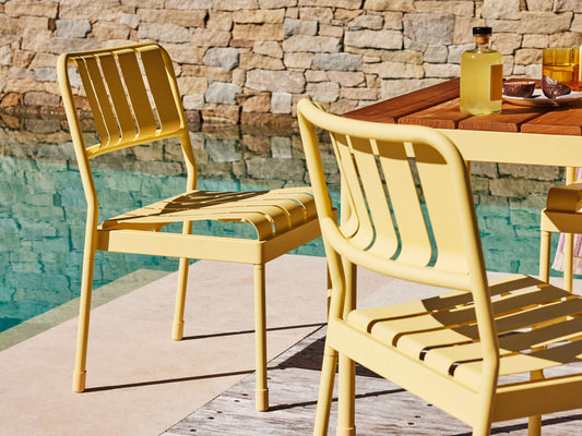 Yellow metal chairs by a wooden table next to a pool. Stone wall in the background. Sunny outdoor setting.