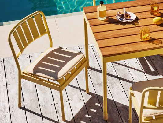 Yellow outdoor chair with a cream cushion and a wooden table on a deck by a pool. Plates and drinks are on the table.