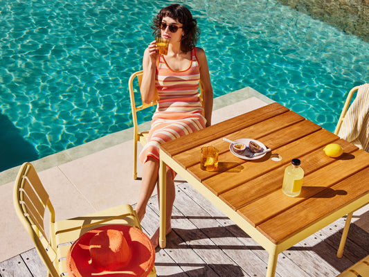 Woman in a striped dress sits at a wooden outdoor table by a pool. Table holds lemons, drinks, and pastries. Bright yellow chairs surround the table.