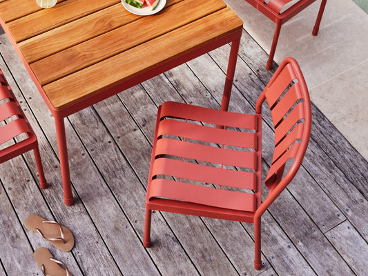 Outdoor setting with a wooden table and red metal chairs on a timber deck. A plate of fruit and thongs are visible.