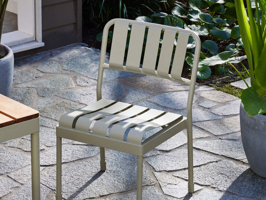 Outdoor chair with light grey metal slats on a stone patio, surrounded by greenery in pots. Ideal for garden or patio furniture settings.