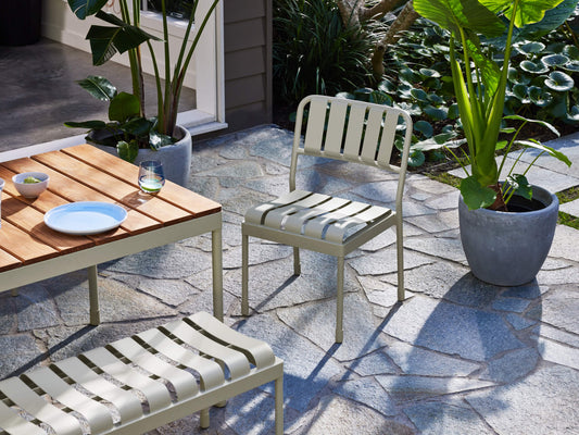 Outdoor patio with light grey metal furniture, a wooden table, and large potted plants on grey stone tiles beside a pond.